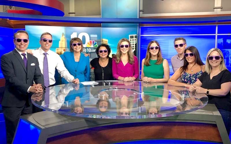 People with sunglasses smiling and standing around a news anchor desk.